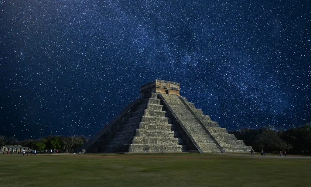 The Chichen Itza pyramid under a star-filled night sky with the Milky Way visible above.